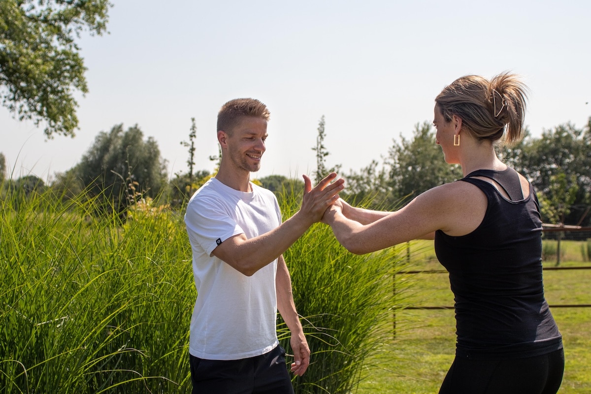 Een man en een vrouw, beiden casual gekleed, staan buiten op een zonnige dag samen een handoefening te doen, glimlachend, met hoog gras en bomen op de achtergrond.