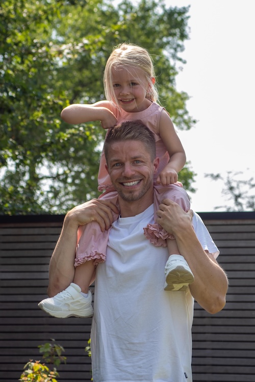 Een lachende man in een wit t-shirt draagt een gelukkig jong meisje op zijn schouders naar buiten, genietend van bewegen zonder pijn. Groene bomen en een houten hek omlijsten het tafereel terwijl het meisje, in een roze outfit en witte gympen, straalt van plezier.