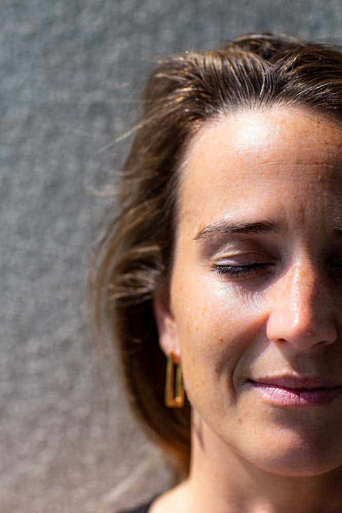 Close-up van een vrouw met lichtbruin haar en gouden oorbellen, de ogen gesloten terwijl het zonlicht haar gezicht belicht tegen een grijze achtergrond met textuur. Dit geeft een sereen moment weer dat actief herstel en gezondheidsbegeleiding op maat weerspiegelt.