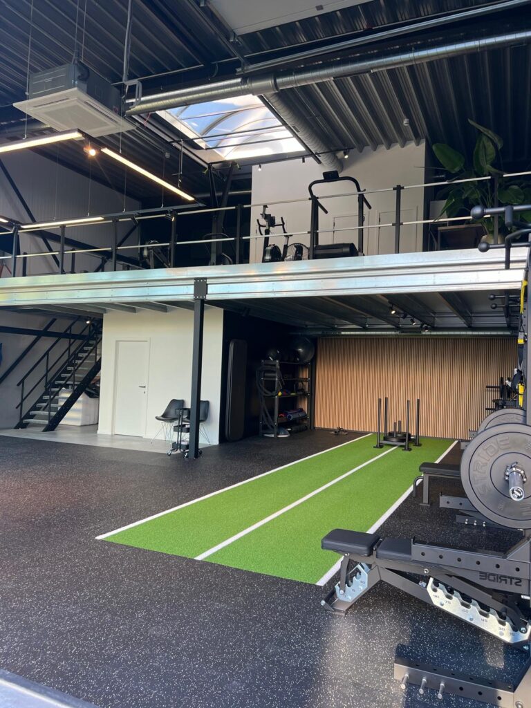 Een moderne fitnessruimte met zwarte vloeren, een groene indoor sprintbaan en cardiotoestellen op een mezzanine. Geniet van een persoonlijk beweegtraject en een industriële inrichting, grote ramen en een trap naar de bovenverdieping.