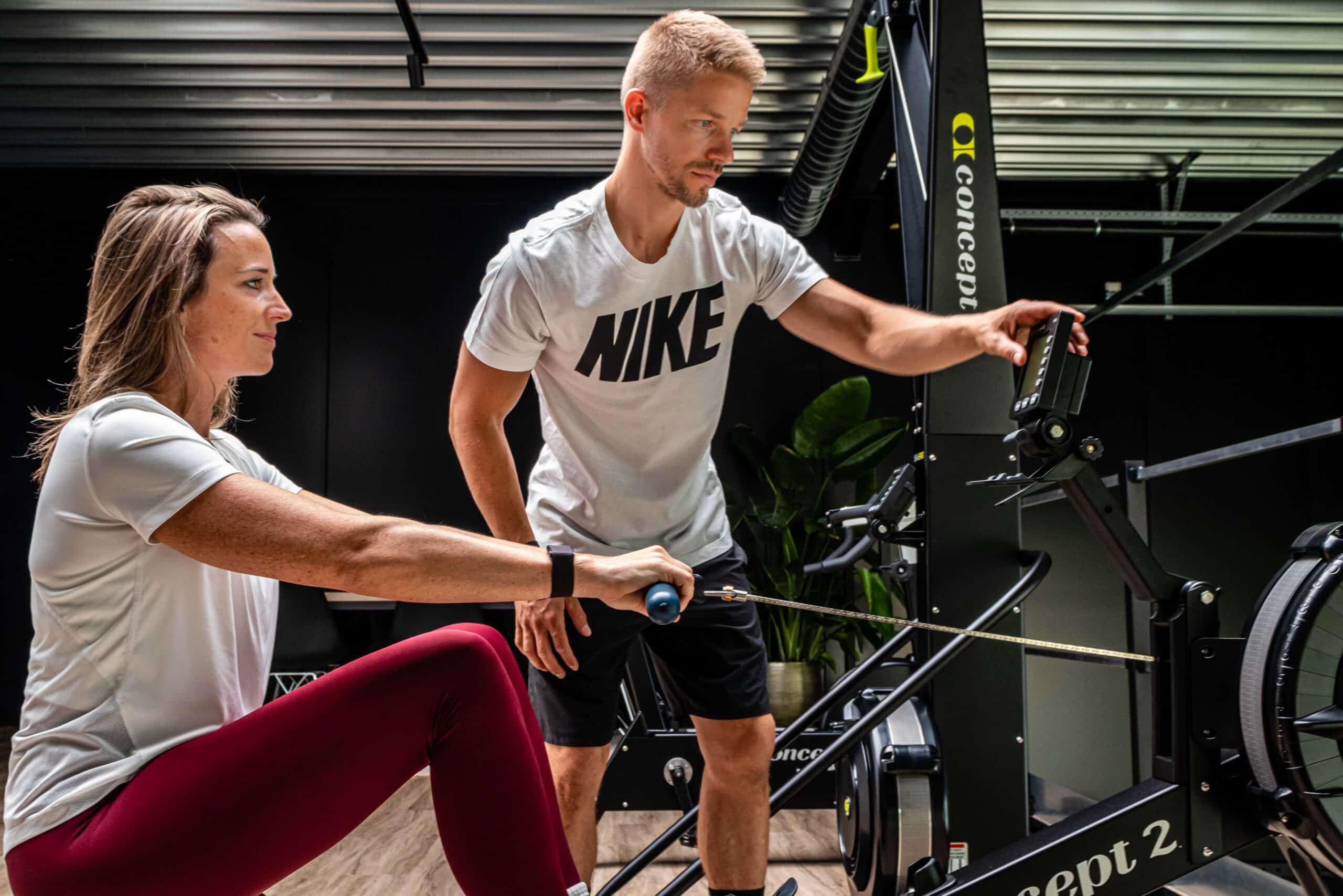 Een vrouw gebruikt een roeimachine terwijl een man in een Nike shirt naast haar staat, de instellingen aanpast en de voordelen intervaltraining uitlegt in een moderne sportschool.