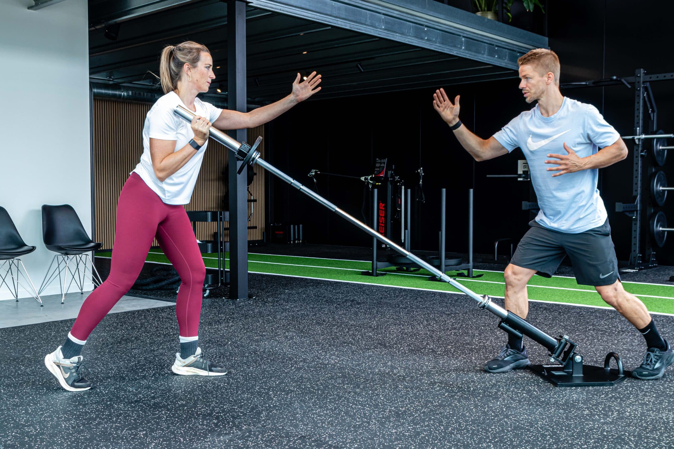Een vrouw voert een landmine press uit met een halter terwijl een man persoonlijke coaching geeft in een moderne sportschool. Beiden doen aan functionele training op een zwarte vloer met apparatuur op de achtergrond.