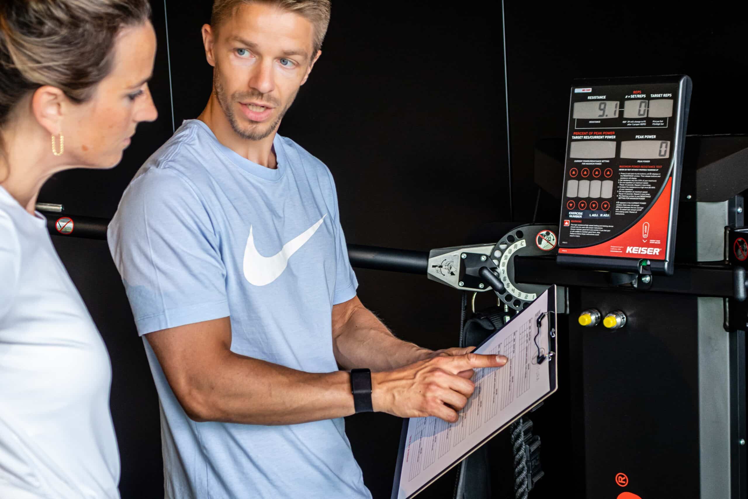 Een man in een lichtblauw shirt toont en legt een flexibel trainingsprogramma op maat uit op een klembord aan een vrouw in een wit shirt in een sportschool, staand naast een krachttrainingstoestel met een digitaal display.