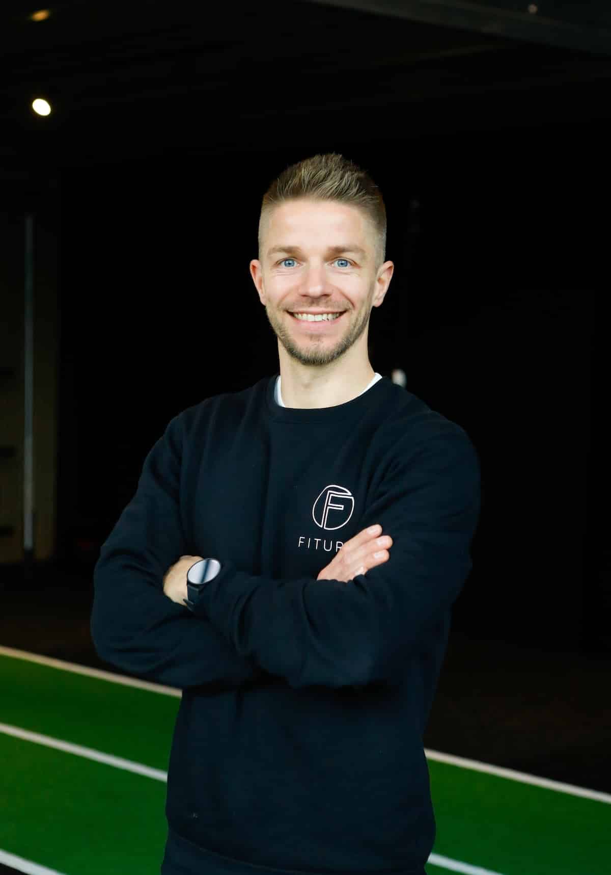 Een lachende man met kort blond haar en een baard staat binnen met zijn armen over elkaar, draagt een zwart sweatshirt met "FITU" en een cirkelvormig logo, voor een groene atletiekbaan.