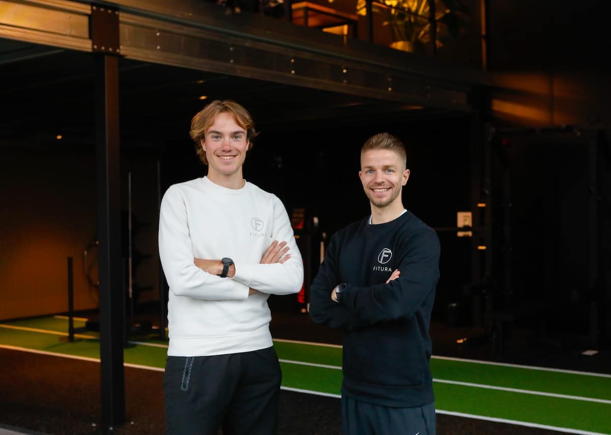 Twee jonge mannen staan zij aan zij in een moderne sportzaal en glimlachen naar de camera. Ze dragen sportkleding met een cirkelvormig logo op de borst. De sportschool heeft kunstgras en zwarte trainingsapparatuur op de achtergrond.
