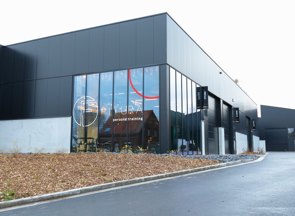 Een modern zwart gebouw met grote glazen ramen met het logo en de naam "Futura Personal Training", omzoomd door een verharde oprit en een aangelegd terrein met mulch.